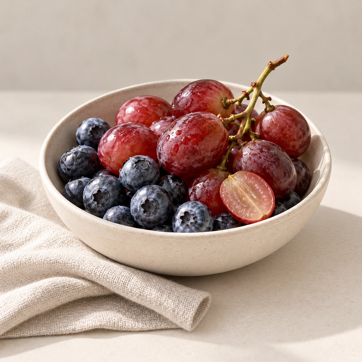 Rote Trauben und Heidelbeeren in einer hellen Keramikschale neben einem hellen Tuch vor neutralem Hintergrund.